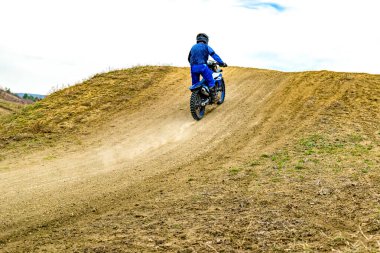 Kask takan bir motokros meraklısı motosikletini toprak bir yolda yarıştırıyor. Açık havada hızla giderken lastikleriyle toz kaldırıyor.