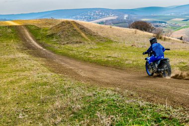 Kişi toprak bir yolda arazi motoruyla manevra yapıyor, otlak yamaçlarla dağlık arazide geziniyor. Aracı engebeli arazide sürerken güvenlik için kask takıyorlar.