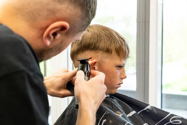 Sarışın ve çilli bir çocuk berber dükkanında bir sandalyede oturur ve sahada gerçek bir profesyonel tarafından kırpılır. Çocuk yeni bir değirmen tarzınız olduğu için çok mutlu. Yüksek kalite fotoğraf