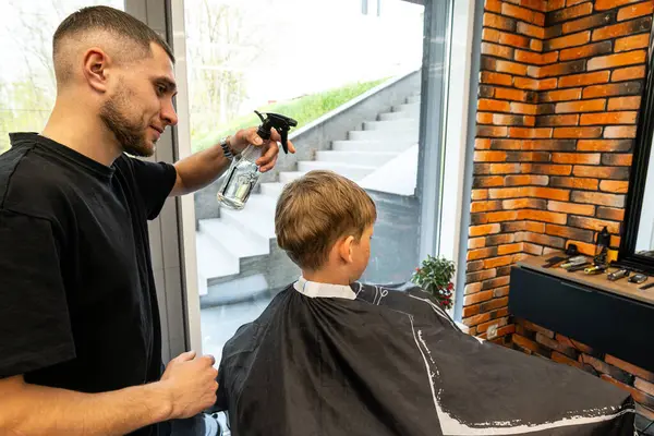 stock image A boy sits in a barbershop while he is sprayed with water to start a haircut. A master in a barbershop sprays water on a boy who is sitting and waiting for his haircut. . High quality photo