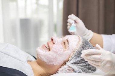 Güzellik salonunda, gülümseyen genç bir kadının portresi. Yüzüne beyaz eldiven giyen bir kozmetik uzmanı tarafından spatulayla kilden bir maske taktıran bir müşteri. Yüksek