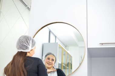 Güzellik Konsepti. Bir güzellik kliniğinde aynaya bakan çekici mutlu bir kadının portresi, güzel bir milenyum bayanı, siyah uzun kollu, yansıması için gülümseyen, görünüşünün tadını çıkaran. Yüksek