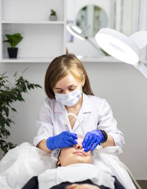 Mikro cilt bakımı uzmanı güzellik kliniğinde gülümseyen bir kadının yüzünü tedavi ediyor. Dermatolog tıbbi eldiven ve yüz maskesi takıyor. Yüksek kalite fotoğraf