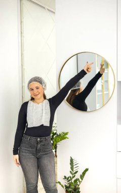 Bir güzellik salonunda, tek kullanımlık bir şapkayla duvara monte edilmiş aynanın önünde gülümseyen ve bir elini havaya kaldıran güzel bir kadının portresi. Yüksek kalite fotoğraf