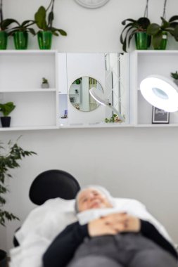 Genç bir kadın ameliyat masasına uzanıyor ve estetik ameliyattan önce ya da sonra gülümsüyor. Çember lamba işyerini aydınlatıyor. Yüksek kalite fotoğraf