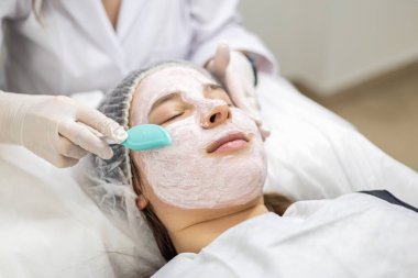 Genç, güzel bir kadın yüzüne beyaz eldiven giyen bir kozmetolog tarafından spatulayla kilden bir maske taktırıyor. Yüksek kalite fotoğraf