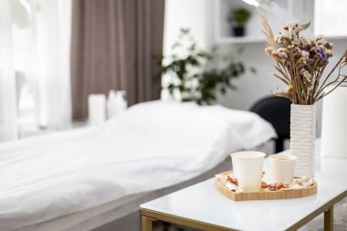 Modern, minimalist güzellik salonu iç mimarisi, beyaz bir masa bir vazo kurutulmuş çiçek, üç fincan kahve veya çay ve biraz şeker sergiler. Arka planda bir prosedür masası görülebilir. Yüksek kalite fotoğraf