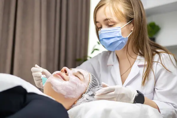 Beyaz eldivenler ve cerrahi maske takan genç bir kozmetik uzmanının yan profil portresi. Müşterinin yüzüne spatula sürerek maske takıyor. Yüksek kalite fotoğraf