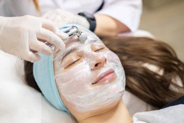 Yüzünde kilden bir maske olan ve yüz masajı yaptıran genç bir kadının yakın çekimi. Kadın güzellik salonunda uzanıyor, yüz bakımı yaptırıyor. Yüzü bir şeyle kaplıydı.