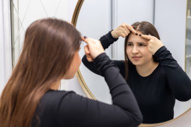 Siyah balıkçı yakalı genç bir kadın, hoşnutsuz görünüyor, cildini inceliyor ve güzellik salonundaki aynaya bakarken güzellik tedavisinden önce durumunu analiz ediyor. Yüksek kalite fotoğraf