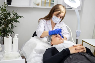 Kozmetik uzmanı müşterilerin yüzleştiği dermabrasionon prosedürünü gerçekleştirir. Uzman beyaz bir takım elbise, yüz maskesi ve tıbbi eldiven giyiyor. Yatay fotoğraf. Yüksek kalite fotoğraf