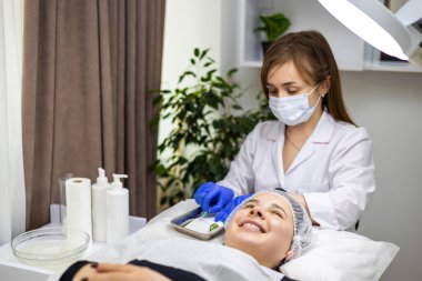 Güzellik kliniğinde tedavi masasına uzanmış genç bir kadının portresi ve cildiyeci, güzellik tedavisi için aletlerini, şırıngasını hazırlıyor. Cilt bakımı kavramı, kozmetoloji. Yüksek kalite