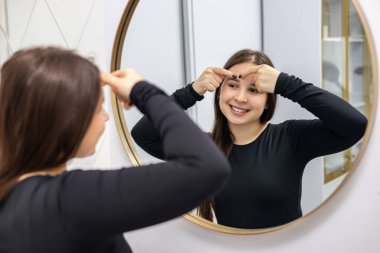 Happy young woman with long hair and a smile on her face looking in the mirror and popping a pimple on her forehead. High quality photo clipart