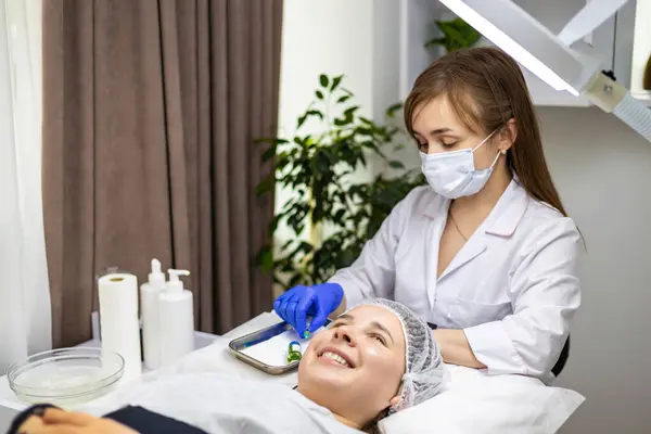 stock image Young experienced dermatologist preparing the syringe and bottle of solution for the following chemical peel, beauty treatment, while her client is lying on a treatment table and smiling. High quality