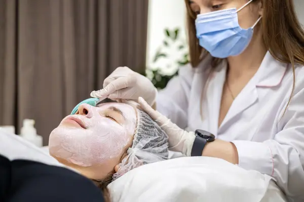Cerrahi maske takan genç bir kozmetolojistin portresi ve beyaz lateks eldivenler, genç bir kadının yüzüne spatulayla yüz maskesi takıyor. Yüksek kalite fotoğraf