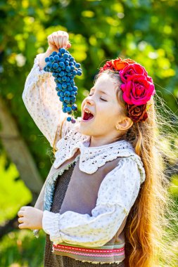 Happy girl eating grapes standing near refrigerator with fruits and vegetables. High quality photo clipart