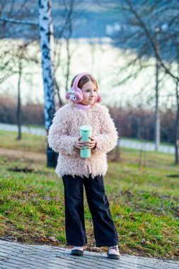 Bahar arka planında çok çekici giyinmiş küçük ve çok tatlı bir kızla çekilmiş bir resim. Küçük kız şık ve elinde insan için çok iyi olan bir termos çay tutuyor.