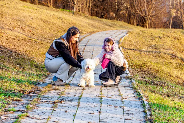 Bahar arka planında, doğanın kucağında çok renkli ve güzel renklerle çekilmiş bir resim. İki kız en iyi arkadaşları olan küçük beyaz köpekle oynuyorlardı.