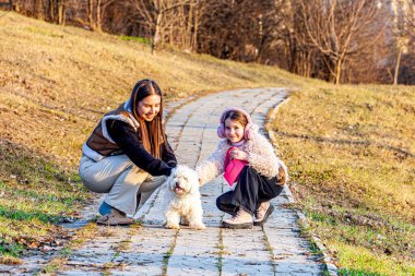 Bahar arka planında, doğanın kucağında çok renkli ve güzel renklerle çekilmiş bir resim. İki kız en iyi arkadaşları olan küçük beyaz köpekle oynuyorlardı.