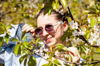 Çiçek açan kiraz ağaçlarının arasında, güneş gözlüğü ve şık bir ceket takan bir kadın, bir bahar öğleden sonrası güneşinin tadını çıkarırken neşeli bir şekilde duruyor. Yüksek kalite fotoğraf