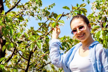 Kiraz bahçesindeyken çok güzel bir genç bayanın fotoğrafı ve çok güzel gülümsüyor. Kız mavi gömlekli ve gözlüklü. Kız çiçek açan meyve bahçesinde poz veriyor.