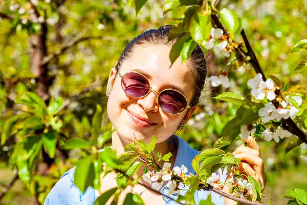 Güneşli bir günde gülümseyen ve çiçek açan elma ağacının kokusunun tadını çıkaran güzel genç bir kadının açık hava portresi. Çiçekli elma bahçesi. Şık bir tişört ve kolye giyiyor. Bahar geldi. Yüksek