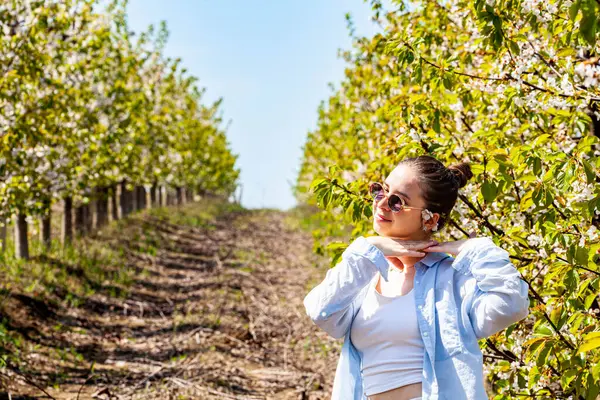 Baharda çiçek açan meyve bahçesinde panoramik manzara. Kiraz bahçesinin manzarası. Hasır şapkalı ve sırt çantalı bir yürüyüşçü kadın baharın tadını çıkarıyor. Yüksek kalite fotoğraf