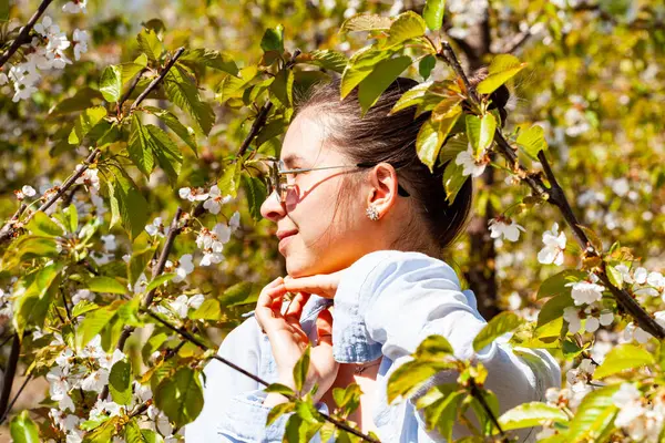Çiçeklerle dolu bir kiraz bahçesinde poz veren çok çekici ve fotojenik genç bir bayanın resmi. Hava güneşli ve çok aydınlık. Meyve bahçesi çok bakımlı ve yeşildir. Yüksek