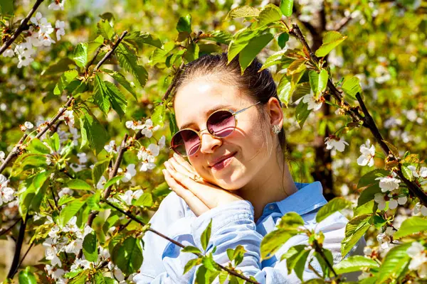 Çiçeklerle dolu bir kiraz bahçesinde poz veren çok çekici ve fotojenik genç bir bayanın resmi. Hava güneşli ve çok aydınlık. Meyve bahçesi çok bakımlı ve yeşildir. Yüksek
