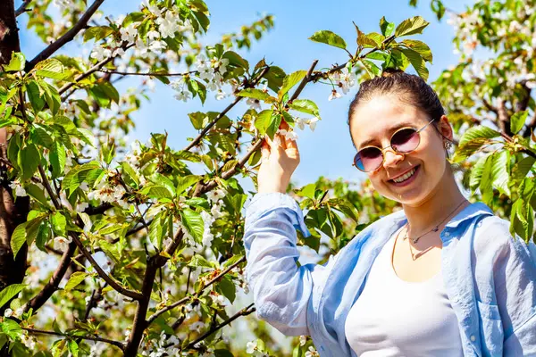 Kiraz bahçesindeyken çok güzel bir genç bayanın fotoğrafı ve çok güzel gülümsüyor. Kız mavi gömlekli ve gözlüklü. Kız çiçek açan meyve bahçesinde poz veriyor.