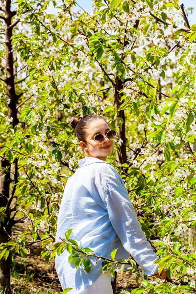 Kiraz bahçesindeyken çok güzel bir genç bayanın resmi ve çok güzel gülümsüyor. Kızın yakalı mavi bir gömleği var. Kız çiçek açan meyve bahçesinde poz veriyor.