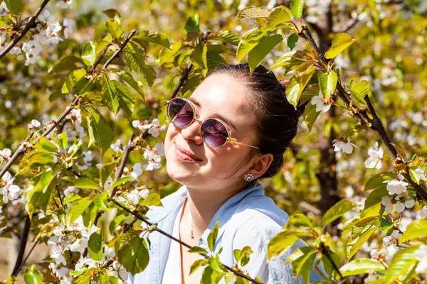 Çiçeklerle dolu bir kiraz bahçesinde poz veren çok çekici ve fotojenik genç bir bayanın resmi. Hava güneşli ve çok aydınlık. Meyve bahçesi çok bakımlı ve yeşildir. Yüksek