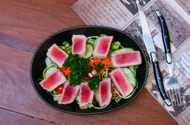 Fresh tuna salad served with sunflower sprouts and fresh organic lettuce in an elegant black plate placed on a wooden table and old newspapers.