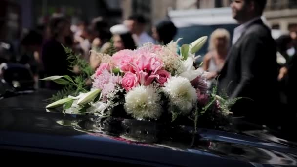ブラックカーのウェディングブーケ 閉じる レトロウェディングカーに置かれた白とピンクのバラの美しいブライダルブーケ バラの花束は黒い自動車の上にあります バックグラウンドに輝くゲスト — ストック動画