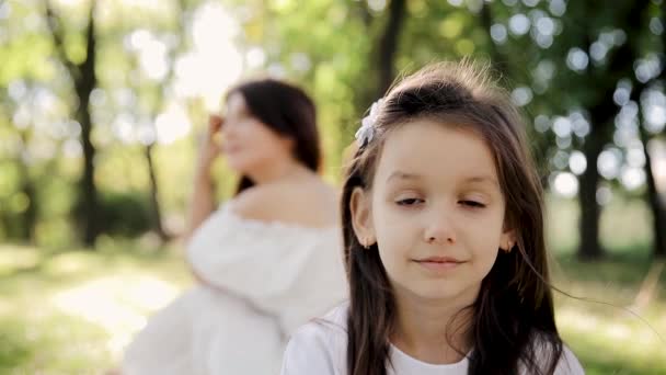 太陽の光の中の子供の幸せな顔 前景にいる少女の肖像画と公園のぼやけた背景にある母親の肖像画 — ストック動画