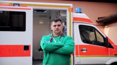 Boynunda steteskop olan beyaz erkek doktor kamera önünde ambulans arabasıyla poz veriyor. Şehir caddesinde duran yeşil üniformalı uzman sağlık görevlisi.