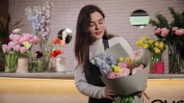 Siyah önlüklü kadın çiçekçi masanın yanında duruyor ve elinde çiçekçide çalışırken bir buket taze çiçek tutuyor.