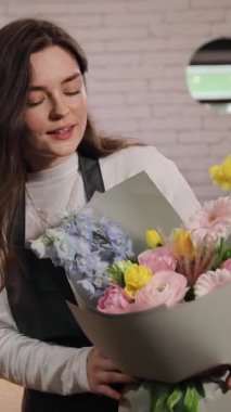 Masanın yanında duran siyah önlüklü kadın çiçekçi çiçekçide çalışırken elinde bir buket taze çiçek tutuyordu. Dikey video