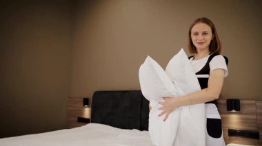 Kendine güvenen otel çalışanı, misafirperverlik ve profesyonelliği vurgulayarak rahat ve temiz bir misafir odasında taze yastıklar sunuyor. Konaklama ve konforlu konaklama