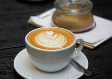 Beyaz kahve fincanı, rosetta latte sanat ve çay tabağı, arka planda kahverengi şekerli şeker kasesi, koyu ahşap masa. Lokantada servis edilen kahveyi kapat..