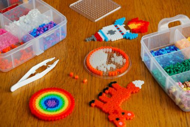 Colorful iron beads (perler beads or fused beads) in a box, art toys created from them and white tweezers on wooden background. Toy developing children's imagination. Concept of children's creativity.