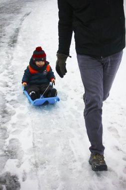 Babası, beyaz oğluyla kızağı karlı yolda çekiyordu. Karlı kış yürüyüşüne çıkmış şirin bir çocuk ve baba. Aile eğlencesi, mutlu bir çocukluk ve kışın açık havada etkinlik..