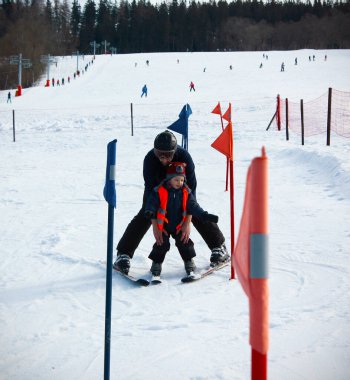 Kurnaz baba, 4 yaşındaki oğluna bebek parkında kayak yapmayı öğretiyor. Örgü şapkalı ve uyarı yeleği giyen küçük kayakçı babasıyla kayak okulunda slalom yapmaya gidiyor..