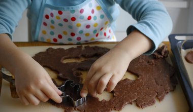 Önlüklü 4 yaşındaki beyaz bir çocuğun elleri rulo zencefilli ekmeğin farklı şekillerini Noel metal kesicilerle kesiyor. Geleneksel Noel kurabiyeleri yapma süreci.