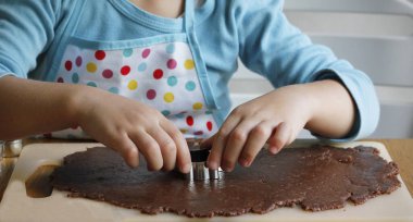 Önlüklü 4 yaşındaki beyaz bir çocuğun elleri rulo zencefilli ekmeğin farklı şekillerini Noel metal kesicilerle kesiyor. Geleneksel Noel kurabiyeleri yapma süreci.