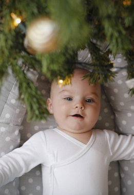 Süslü bir Noel ağacının altında bebek yuvasında yatan beyaz elbiseli, sevimli ve neşeli beş aylık beyaz bir bebek. Bebeğin ilk Noel 'i. Üst görünüm.