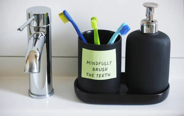 Close-up of a water tap, a black soap dispenser and a cup for toothbrushes with a sticker containing a handwritten inscription: Mindfully brush the teeth. Mindfulness training concept
