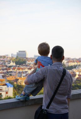 Kareli gömlek giyen beyaz bir baba 3 yaşındaki oğluna sarılıyor ve akşam güneşinde Prag 'ın manzarasına bakıyorlar. Arkadan vurulmuş. Kopya alanlı Babalar Günü konsepti.