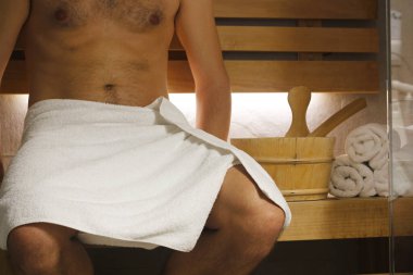 Kalçasının üzerinde beyaz bir havlu olan yarı çıplak bir adamın yakın çekimi saunadaki ahşap bir bankta, yanında kepçeli sauna kovası ve sarılmış beyaz havlular, arka ışıklandırmayla aydınlatılmış sauna..