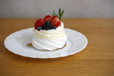 Beyaz bir tabakta servis edilen taze böğürtlenlerle süslenmiş mini pavlova pastası, arka planda tahta masa ve gri duvar. Seçici odak, boşluğu kopyala